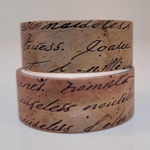 Pode incluir: Dois rolos de fita washi decorativa. A fita tem uma aparência vintage e envelhecida com um esquema de cores castanho e bege. A fita é coberta por escrita manuscrita em tinta preta, adicionando à sua estética antiga.
