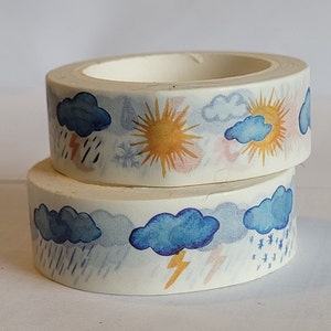 Op de afbeelding: Twee rollen witte washi tape met een herhalend patroon van blauwe wolken, gele zonnen en regendruppels. De tape is versierd met een aquarel-achtig ontwerp.