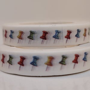 May include: Two rolls of white washi tape with a repeating pattern of colorful push pins. The push pins are red, yellow, green, and blue.