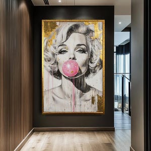 Op de afbeelding: Een ingelijst portret van een vrouw die een roze kauwgomballenblaas blaast. Het kunstwerk heeft een gouden lijst en accenten van bladgoud. De vrouw heeft blond haar en draagt dramatische make-up. Roze verf druipt van de bel af.