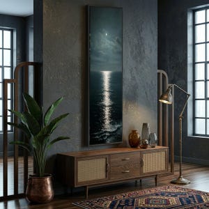 May include: A framed vertical art print depicts a moonlit ocean scene with a bright, reflective path of light. The artwork hangs above a wooden cabinet with woven panels, complemented by decorative vases, a floor lamp, and a patterned rug. A large potted plant adds a touch of nature.