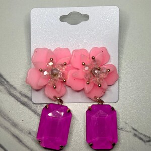 Puede incluir: Un par de pendientes de flores rosas con centros de cuentas transparentes y detalles dorados. Cada pendiente tiene un colgante de gema rectangular grande de color magenta. Los pendientes se muestran en una tarjeta blanca.