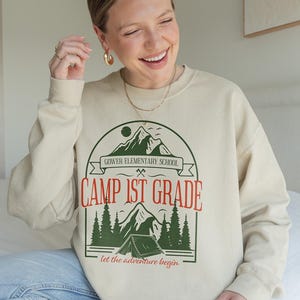 May include: A beige sweatshirt featuring a green and red graphic with the text "Camp 1st Grade" and a mountain and tent design. The phrase "let the adventure begin" is below the graphic. The sweatshirt is worn by a person.