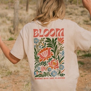 Puede incluir: Camiseta beige con un dise&ntilde;o floral y la palabra "BLOOM" en letras rojas. El dise&ntilde;o floral incluye flores y hojas rosas, naranjas, azules y verdes. El texto "WHERE YOU ARE PLANTED" est&aacute; debajo del dise&ntilde;o floral.