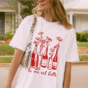 Peut inclure: T-shirt blanc avec un motif rouge de bouteilles de vin avec des fleurs &agrave; l'int&eacute;rieur. Le texte "la vie est belle" est imprim&eacute; sous le motif.