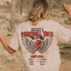 May include: A beige t-shirt with a graphic of wings and a lightning bolt. The text "Mecan's Farewell Tour May 2024" is printed on the shirt. The text "Guest Stars" is printed below the graphic, followed by a list of names.