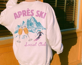 Op de afbeelding: Wit sweatshirt met een grafische print van een winterscene met bergen, skiërs en champagne glazen. De tekst "APRÉS SKI" en "Social Club" staat op het sweatshirt gedrukt.