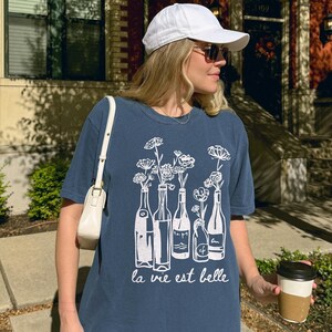 Peut inclure: Une femme portant un t-shirt bleu avec un motif blanc de bouteilles de vin et de fleurs. Le t-shirt dit "la vie est belle". Elle porte une casquette de baseball blanche et des lunettes de soleil. Elle tient une tasse de caf&eacute;.
