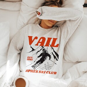 May include: A gray sweatshirt with a red and black graphic print of a skier in the mountains. The text "Vail" is printed in red above the graphic and "Est. 1881" and "Apres Ski Club" are printed in black below the graphic.