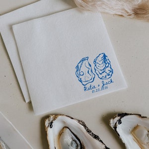 May include: Two white oyster shells on a white surface with a white napkin with a blue oyster illustration and the text "Katie + Jack 01.22.2025" printed on it.