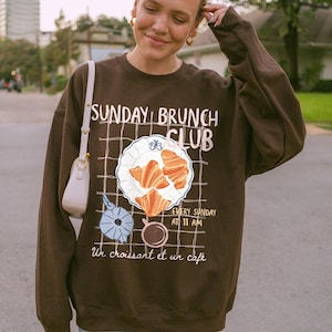 May include: A brown sweatshirt with a graphic print of a croissant and coffee cup. The text on the sweatshirt reads "Sunday Brunch Club" and "Un croissant et un cafe".