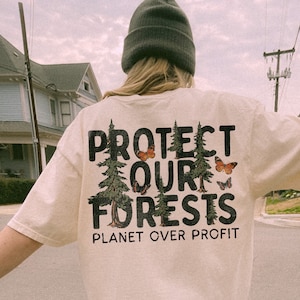 May include: A person wearing a light beige t-shirt with the text "Protect Our Forests, Planet Over Profit" printed on the back. The shirt features a graphic of trees and butterflies.