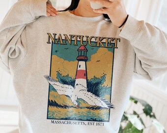Könnte beinhalten: Hellgraues Sweatshirt mit einem Grafikdruck. Der Druck zeigt einen Leuchtturm, eine Möwe und Meereswellen, mit dem Wort "Nantucket" darüber und "Massachusetts, EST 1671" darunter.