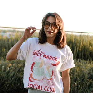 May include: A white t-shirt with a graphic of a cocktail drink and the text "extra spicy margs social club". The person wearing the shirt is also wearing blue jeans and sunglasses.