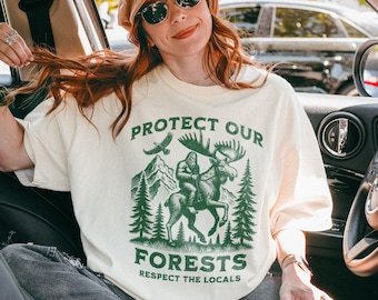 Pode incluir: Uma mulher usando um chapéu bege e óculos de sol está sentada em um carro. Ela está vestindo uma camiseta creme com um gráfico verde que diz "Proteja nossas florestas Respeite os moradores locais". O gráfico apresenta um Pé Grande montando um alce por uma floresta.