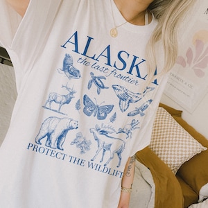 Puede incluir: Camiseta blanca de gran tama&ntilde;o con un estampado gr&aacute;fico azul de varios animales salvajes de Alaska, incluyendo un oso, una ballena, una mariposa y un alce. El texto "Alaska the last frontier" y "Protect the wildlife" est&aacute; impreso en azul en la camiseta.