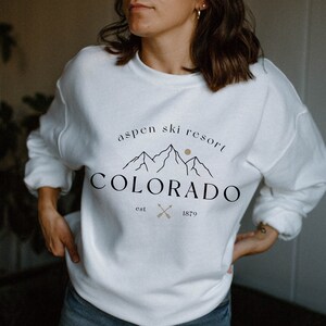 K&ouml;nnte beinhalten: Wei&szlig;es Sweatshirt mit dem Text "Aspen Ski Resort Colorado" in Schwarz und Beige. Das Design umfasst Berggrafiken, eine Sonne und gekreuzte Pfeile. Das Sweatshirt ist ein l&auml;ssiger Stil.