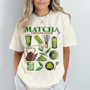 Puede incluir: Una camiseta de color crema con un gráfico verde que dice "Matcha Lovers Club". El gráfico presenta ilustraciones de té matcha, un batidor, una taza de té y una tetera.