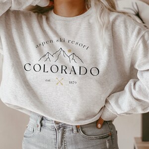 K&ouml;nnte beinhalten: Ein hellgraues Sweatshirt mit dem Text "Aspen Ski Resort Colorado" in Schwarz, mit einer Berg-Grafik und dem Jahr "1879". Das Sweatshirt hat einen Rundhalsausschnitt.