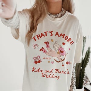K&ouml;nnte beinhalten: Wei&szlig;es T-Shirt mit einer roten und pinkfarbenen Grafik eines Amor mit Pfeil und Bogen. Der Text "That's Amore est. 2025 Katie and Marc's Wedding" ist auf das Shirt gedruckt.