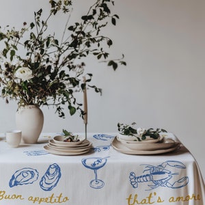 Peut inclure: Nappe blanche avec des illustrations bleues et jaunes de homards, d'huîtres et d'un verre à martini. Le texte "Buon appetito" et "that's amore" est imprimé sur la nappe. Un vase blanc avec des fleurs blanches et une pile d'assiettes blanches sont sur la table.