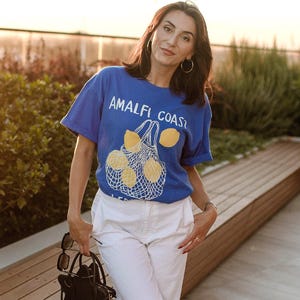 May include: A woman wearing a blue t-shirt with the text "Amalfi Coast" and a lemon graphic, white pants, and black sandals. She is standing on a patio with a wooden bench behind her.