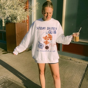 May include: White sweatshirt with a graphic design of a cafe menu. The text on the sweatshirt reads "Sunday Brunch Club" and "Every Sunday at 11 AM" with a croissant and coffee illustration. The text "Un croissant et un cafe" is also included on the sweatshirt.