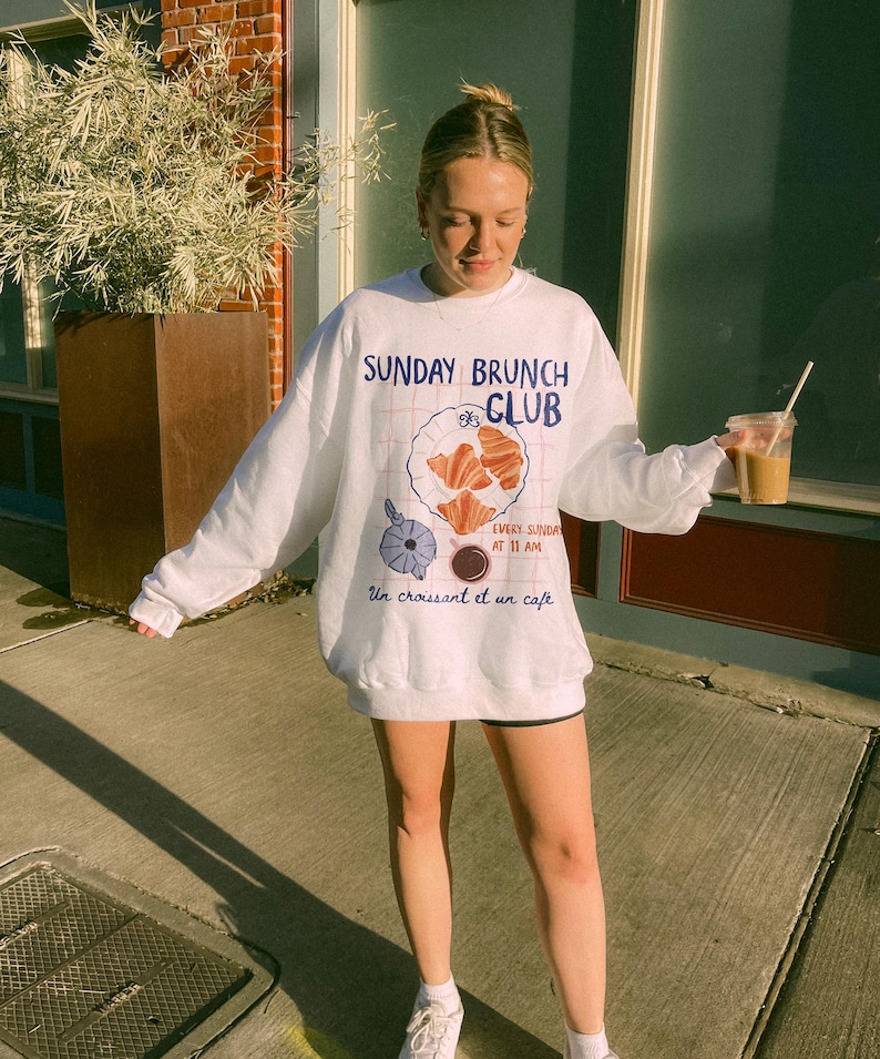 May include: White sweatshirt with a graphic design of a cafe menu. The text on the sweatshirt reads "Sunday Brunch Club" and "Every Sunday at 11 AM" with a croissant and coffee illustration. The text "Un croissant et un cafe" is also included on the sweatshirt.