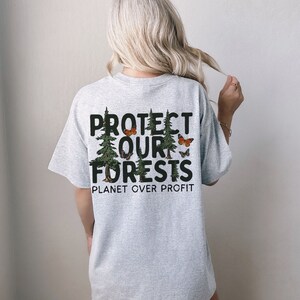 Op de afbeelding: Grijs T-shirt met een grafische print van bomen en vlinders. De tekst op het shirt luidt "Protect Our Forests Planet Over Profit".