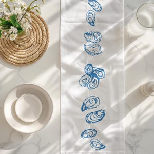 May include: White table runner with a blue oyster design. The runner is on a white marble surface with a bowl and a woven coaster.