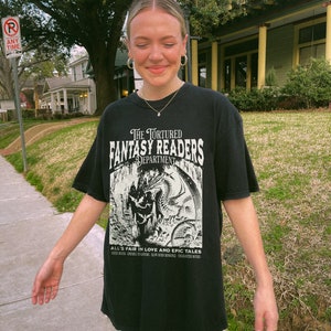 May include: A black t-shirt with a white graphic print of a dragon and the text "The Tortured Fantasy Readers Department" and "All's Fair in Love and Epic Tales".