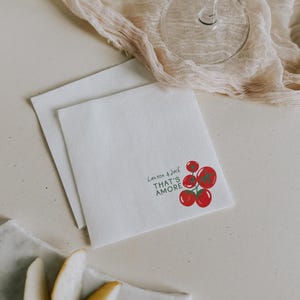 May include: Two white square napkins with the text "Lauren & Jack THAT'S AMORE" and a red tomato illustration. A glass of wine and a beige cloth are in the background. Slices of pear are on a marble plate.