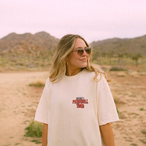 May include: A woman wearing a white t-shirt with the text "Megan's Farewell Tour" printed in red on the front. She is wearing sunglasses and looking to the right. The background is a desert landscape.