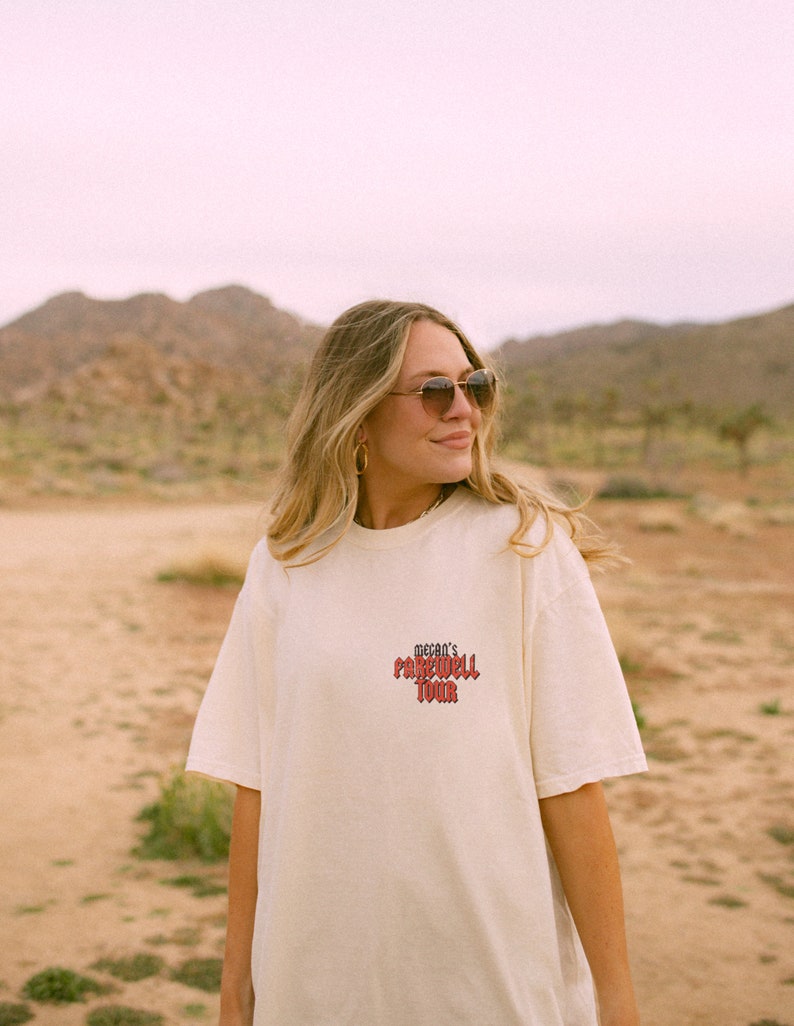 May include: A woman wearing a white t-shirt with the text "Megan's Farewell Tour" printed in red on the front. She is wearing sunglasses and looking to the right. The background is a desert landscape.