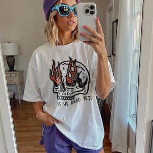 May include: A white t-shirt with a skull graphic and the text "Is he dead yet?" The skull has flames coming out of its eye sockets. The model is wearing a purple beanie and blue sunglasses.