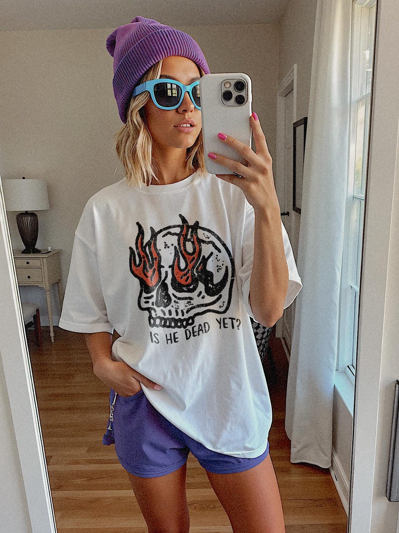May include: A white t-shirt with a skull graphic and the text "Is he dead yet?" The skull has flames coming out of its eye sockets. The model is wearing a purple beanie and blue sunglasses.
