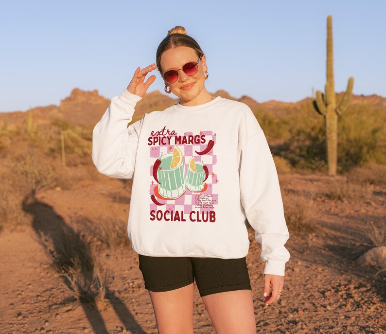 May include: White sweatshirt featuring the text "extra SPICY MARGS SOCIAL CLUB" and a graphic of two cocktails with chilli peppers and lemon slices. The background is a pink and white chequered pattern.