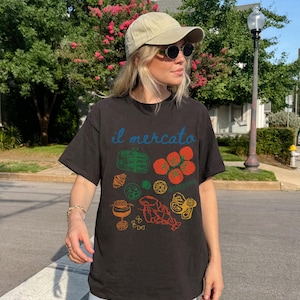 May include: A black t-shirt with a colorful graphic print of various Italian food items, including tomatoes, pasta, and a lobster. The text "il mercato" is printed on the shirt in blue.