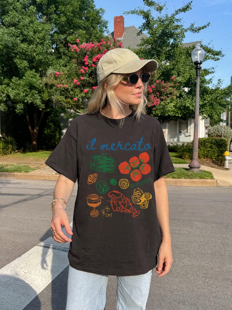 May include: A black t-shirt with a colorful graphic print of various Italian food items, including tomatoes, pasta, and a lobster. The text "il mercato" is printed on the shirt in blue.
