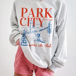 Op de afbeelding: Grijs sweatshirt met een blauwe en rode grafische print met de tekst "Park City Est. 1881 apres ski club".