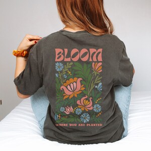 Puede incluir: Camiseta gris oscuro con un dise&ntilde;o floral y la palabra "BLOOM" en letras color coral. El dise&ntilde;o floral incluye flores y hojas rosas, naranjas, azules y verdes. El texto "WHERE YOU ARE PLANTED" est&aacute; debajo.