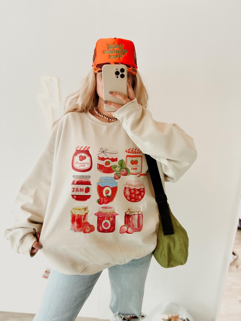 May include: A cream-colored sweatshirt with a graphic print of multiple jars of strawberry jam. The sweatshirt has a crew neck and long sleeves. The model is wearing a bright orange baseball cap with the text "Going Nowhere Fast" in green.
