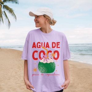 May include: A light purple t-shirt with the words "AGUA DE COCO" in red, and a green coconut illustration with a straw. The text "praia de copacabana" is below the coconut. The shirt is worn by a person on a beach.