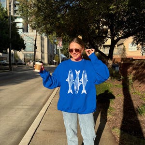 May include: A woman wearing a royal blue sweatshirt with a white graphic of three fish. She is wearing light blue jeans and white sneakers. She is holding a brown to-go coffee cup.