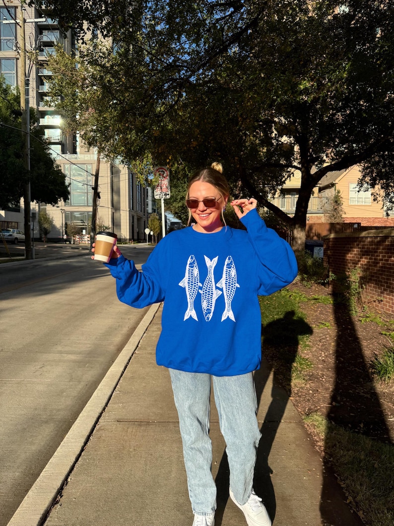 May include: A woman wearing a royal blue sweatshirt with a white graphic of three fish. She is wearing light blue jeans and white sneakers. She is holding a brown to-go coffee cup.