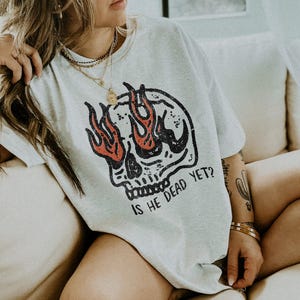 May include: A gray t-shirt with a skull graphic and flames. The skull has a single eye with flames coming out of it. The text "Is he dead yet?" is printed below the skull.