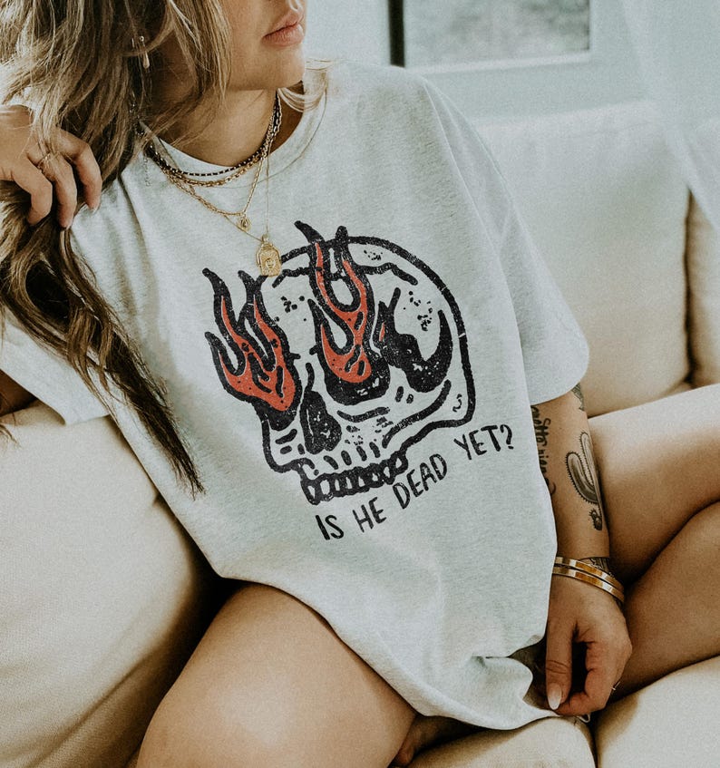 May include: A gray t-shirt with a skull graphic and flames. The skull has a single eye with flames coming out of it. The text "Is he dead yet?" is printed below the skull.