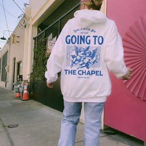 May include: A white hoodie with the text "LOVE IS IN THE AIR EST. 2025 GOING TO THE CHAPEL LAS VEGAS, NV" in blue. The design includes two cherubs. The person is wearing light blue jeans and a pink cap.