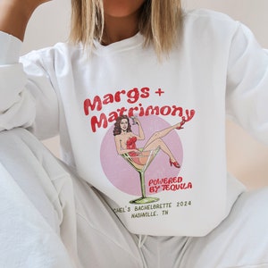 May include: White sweatshirt with a graphic print of a woman in a red dress sitting in a martini glass. The text on the shirt reads "Margs + Matrimony Powered by Tequila Chel's Bachelorette 2024 Nashville, TN".