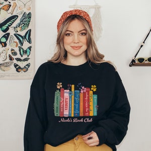 Op de afbeelding: Zwart sweatshirt met een afbeelding van kleurrijke boeken met bloemmotieven en de tekst "Nicole's Book Club".